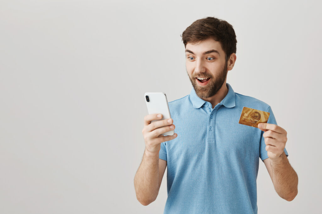 man received cashback for his purchase. portrait of excited and happy bearded european male holding credit card and smartphone, looking at screen with emotive and upbeat expression over gray wall